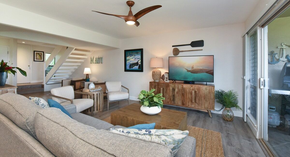 Stylish living room at Kaanapali Plantation 25 with modern gray sofa, wooden coffee table, and TV on a rustic console. Bright, airy vibe with plants and ocean artwork.