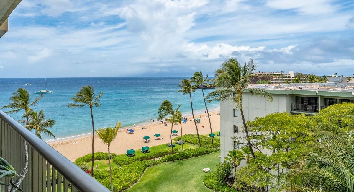 A serene beach scene from Whaler 712 with turquoise waves, palm trees, and colorful umbrellas on golden sand. A balcony overlooks the view, creating a tropical vibe.