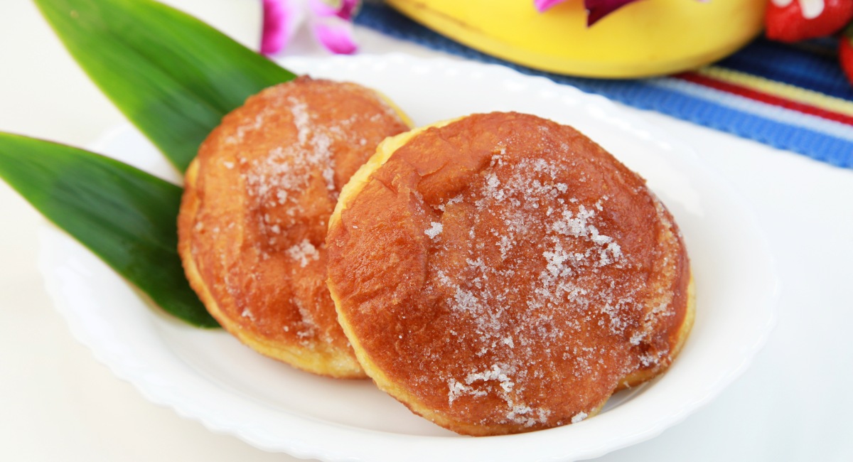 Sugar-dusted malasadas on a white plate with a ti leaf garnish — a specialty at Komoda Store & Bakery, a Makawao institution since 1916
